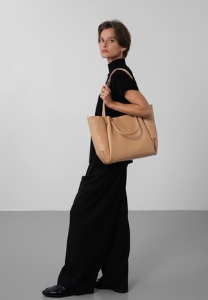 Femme en tenue noire tenant un grand sac fourre-tout en cuir beige sur l'épaule, debout devant un fond gris et blanc uni.