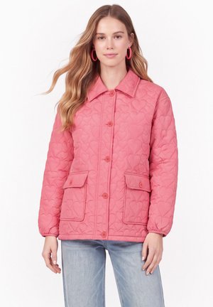 Femme portant une veste matelassée rose avec des motifs en forme de cœur, des fermetures à boutons, des poches à rabat et un jean bleu clair sur fond blanc.
