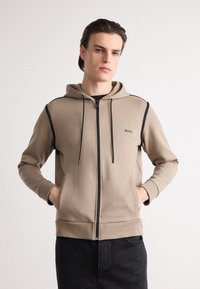 Veste à capuche beige zippée avec des accents noirs et des poches avant. Présente un petit logo "BOSS" sur la poitrine. Tissu lisse de poids moyen.
