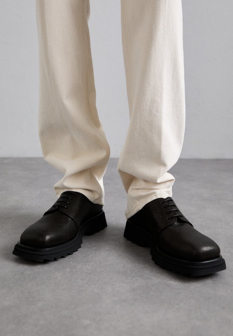 Fekete bőr cipő texturált felülettel, kerek orral és fűzős elején. Gumi talp, amelyen barázdák találhatók a tapadás érdekében, krém színű nadrággal viselve.