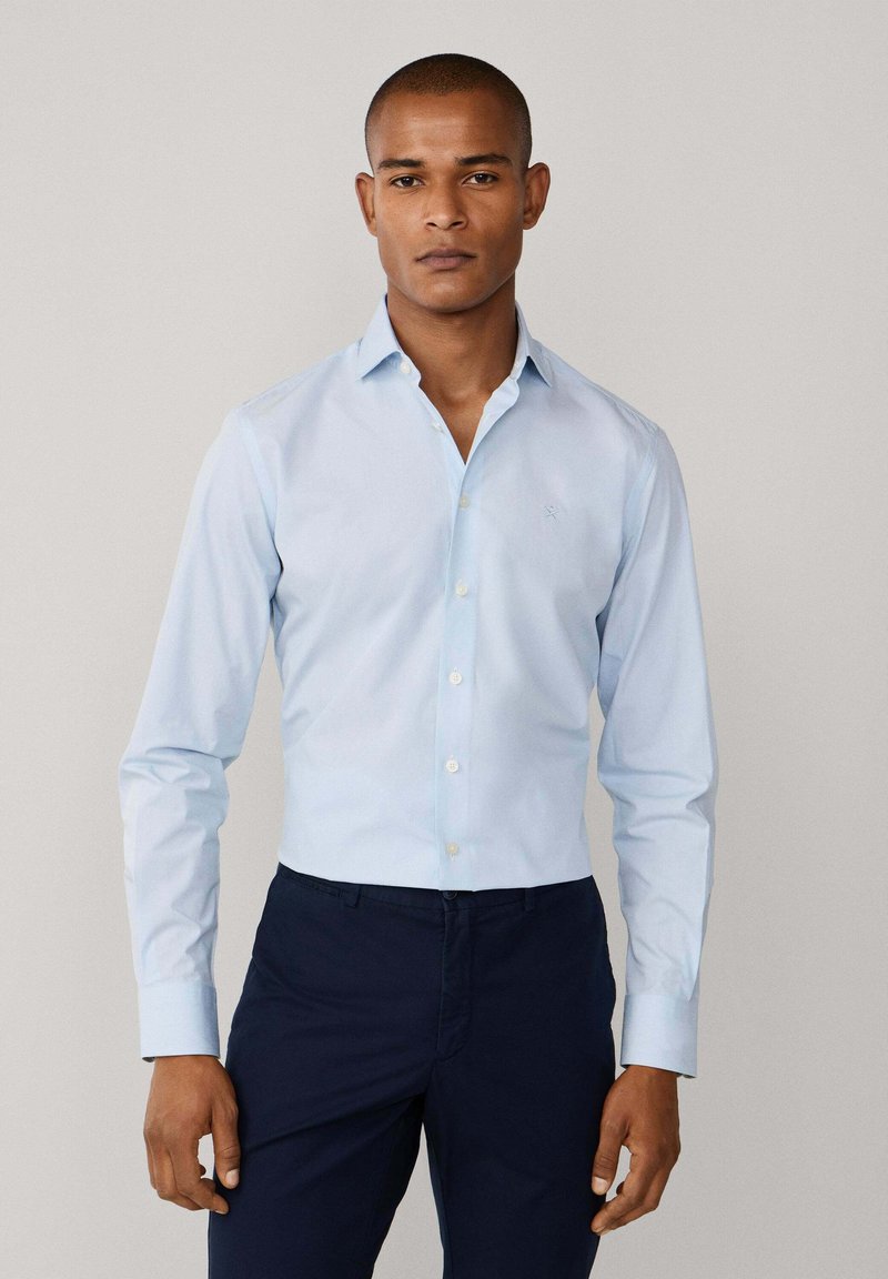 Svijetloplava košulja s dugim rukavima i ovratnikom na gumbe, bijelim gumbima. Ima mali logo na prsima. Tamnoplave hlače.