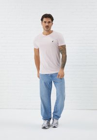Camiseta corta de manga rosa claro con un logo negro en el pecho, combinada con unos jeans de mezclilla azules y zapatillas deportivas grises. De pie sobre un suelo blanco.