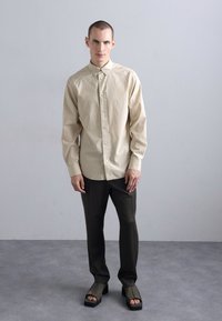 Beige knap-up skjorte, mørkegrønne bukser og sorte sandaler. Skjorten har krave og manchetter; glat tekstur og afslappet pasform.