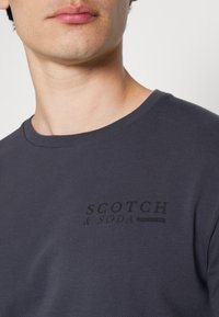 Tmavě šedé tričko s kulatým výstřihem z bavlny, na levé části hrudi s malým černým logem "SCOTCH & SODA".