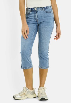 Mujer que lleva vaqueros recortados de mezclilla azul claro, zapatillas blancas y anillos en los dedos, de pie frente a un fondo liso.