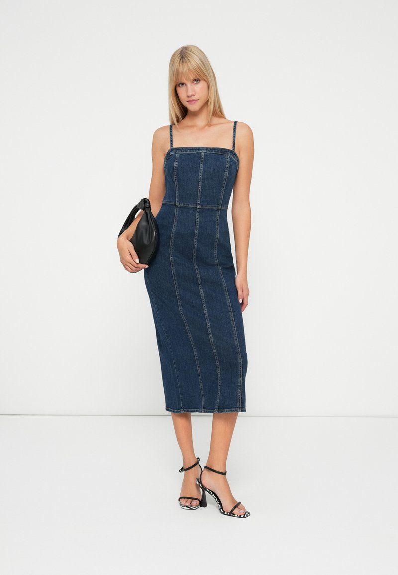 Vrouw in een aansluitende denim midi-jurk met spaghettibandjes, houdt een zwarte handtas vast en draagt zwart-witte gestreepte hakken.