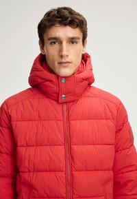 Giacca a vento rossa con collo alto, chiusura a zip e bottoni a pressione. La texture è liscia con motivi trapuntati e tasche laterali.