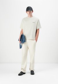 Helles graues T-Shirt mit einem kleinen Druck, beige Hosen, blaue Jeansjacke über dem Arm, schwarze Kappe und schwarze Schuhe.