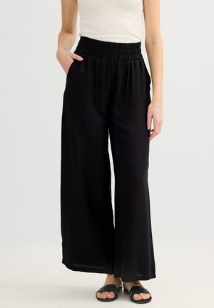 Mujer con pantalones anchos negros de cintura elástica y bolsillos laterales, combinados con sandalias negras y una camiseta blanca sin mangas.