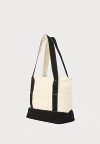 Υφασμάτινη τσάντα tote με κρεμ σώμα και μαύρο κάτω μέρος. Διαθέτει διπλούς μαύρους ιμάντες και λευκή ραφή. Ορθογώνιο σχήμα, ανοιχτό επάνω μέρος.