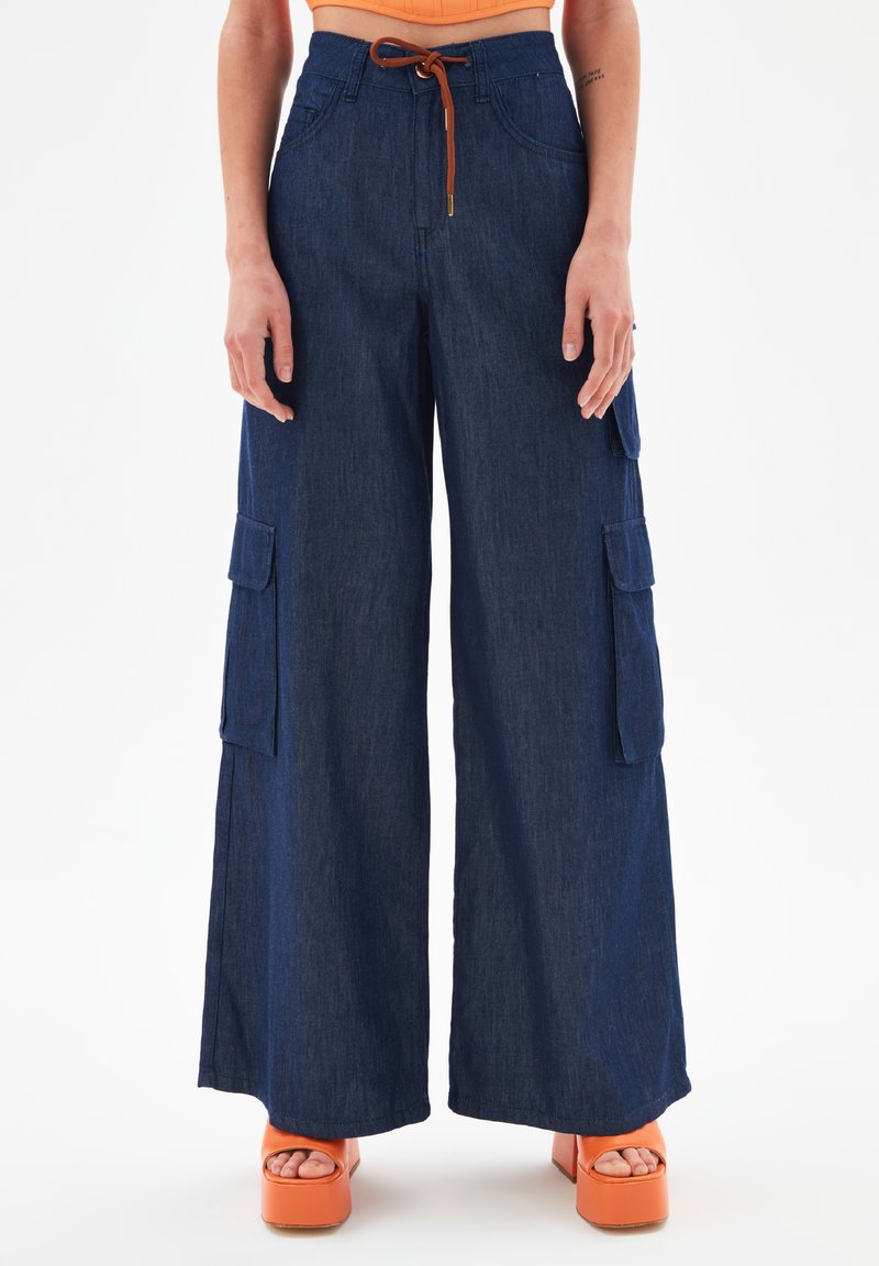 Pantalon en denim foncé à jambes larges avec poches avant et poches cargo latérales, maintenu par une ceinture à cordon marron, porté avec des sandales à plateforme orange.