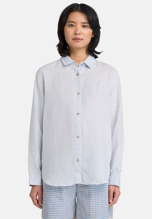 Młoda kobieta o ciemnych włosach do ramion, ubrana w jasnoniebieską koszulę na guziki i niebieskie spodnie w kratę, stojąca na białym tle.