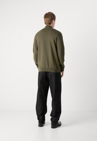 Olivgrüner gestrickter Pullover mit einem gerippten Kragen, langen Ärmeln und einem schwarzen Patch am Ärmel. Getragen mit locker sitzenden dunklen Hosen.
