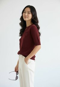 Bordowa bluzka z krótkim rękawem z teksturowanymi ozdobami w kształcie serc, w połączeniu z kremowymi spodniami. Modelka trzyma czarne okulary przeciwsłoneczne, stoi na jasnym tle.