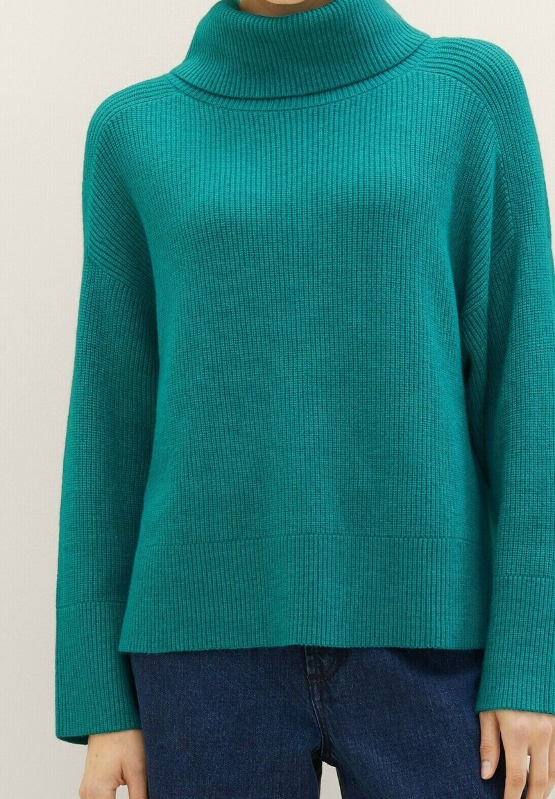 Pull col roulé côtelé couleur sarcelle à manches longues, porté avec un jean bleu foncé, visible des épaules jusqu'aux hanches.