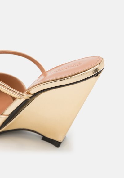 Sandalia cuña dorada con un tacón curvado, tiras finas y una textura suave. Presenta una plantilla de color beige claro y un acento negro en la parte inferior.