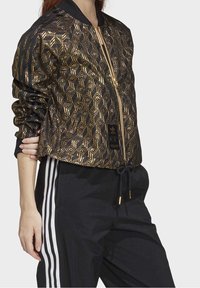 Černá bomber bunda se zlatým geometrickým vzorem, zapínání na zip, žebrovaný límec, elastické manžety a lem, spárovaná s černými tepláky s bílými pruhy.