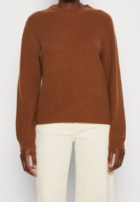 Suéter marrón de punto con cuello redondo y textura acanalada, con mangas largas y un sutil logo en la parte baja delantera. Combinado con pantalones blancos.