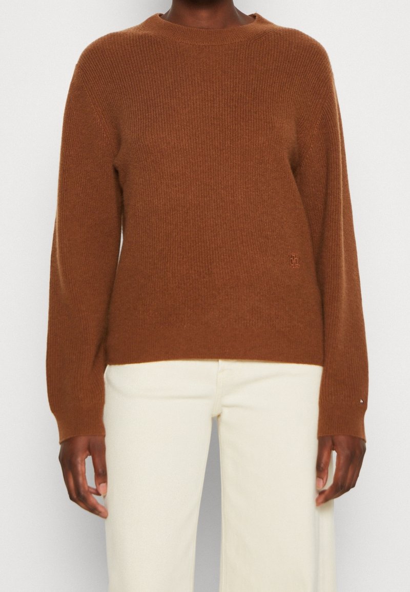 Suéter marrón de punto con cuello redondo y textura acanalada, con mangas largas y un sutil logo en la parte baja delantera. Combinado con pantalones blancos.