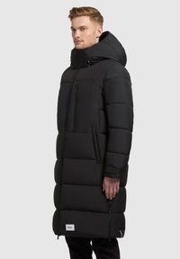 Lang sort pufferjakke med hætte, med quiltede sektioner, lynlåslomme og logo-patch på den nederste kant. Glat, mat tekstur.