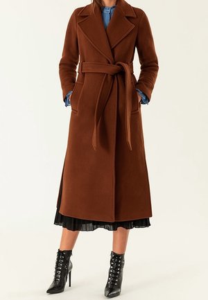 Frau trägt einen langen braunen Mantel mit Gürtel über einem schwarzen, plissierten Rock und schwarzen, geschnürten Stiefeln mit hohem Absatz, Hände in den Manteltaschen.