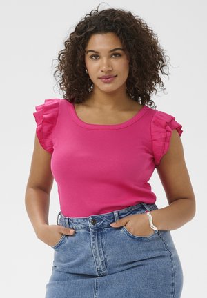 Kvinde med krøllet hår iført en knaldrød, ærmeløs top med flæser ved skuldrene og højtaljede blå jeans, hænderne i lommerne.