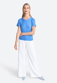 Top corto a maniche corte blu a coste con orlo ondulato, abbinato a pantaloni bianchi a gamba larga e scarpe azzurre a punta.