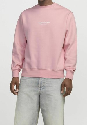 Homme portant un sweat-shirt rose clair avec le texte "Originals Studio" sur la poitrine et un jean ample délavé clair, sur fond blanc uni.