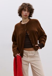 Hnědý pletený kardigan s kulatým výstřihem a zapínáním na knoflíky, doplněný krémovými kalhotami a červeným huňatým šátkem, který zdůrazňuje strukturované materiály.