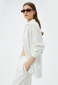 Wit gestreepte shirt met een relaxte pasvorm, doorgeknoopte voorkant en opgerolde mouwen, gecombineerd met witte broek en bruine rechthoekige zonnebril.