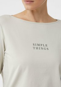 Jasnoszara bluzka z długim rękawem, z szerokim dekoltem i czarnym napisem "SIMPLE THINGS" wyśrodkowanym na klatce piersiowej. Gładka faktura materiału.