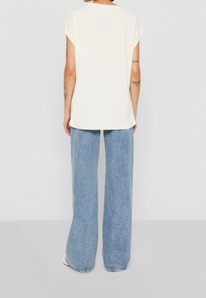 Person vendt væk iført en løs hvid kortærmet top og lyseblå jeans med vide ben, stående på en ensfarvet grå gulv.