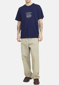 T-shirt en coton bleu marine à manches courtes avec un motif imprimé, associé à un pantalon en denim beige clair et des chaussures marron.