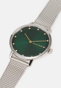 Kerek zöld számlapos óra ezüst szövött pánttal, arany mutatókkal és kristály jelzőkkel. Hilfiger márkázott, és textúrázott számlappal rendelkezik.