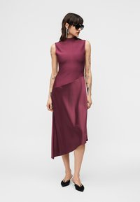 Γυναίκα με τατουάζ που φοράει αμάνικο, ασύμμετρο φόρεμα μπορντό με λαιμόκοψη, μαύρα μυτερά παπούτσια slingback, μαύρα γυαλιά ηλίου και μακριά σκουλαρίκια.