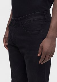 Mann trägt schwarze Jeans und schwarzes Hemd, mit der rechten Hand entspannt an der Seite, auf hellem einfarbigem Hintergrund.
