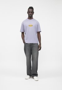 Lavandas krāsas īsroku T-krekls ar dzeltenu logotipu "GUESS", kombinēts ar vaļīgām pelēkām džinsa biksēm un baltām teniskām. Vienkāršs fons.