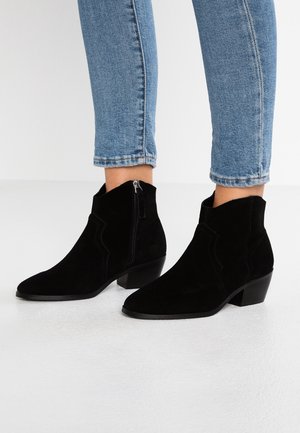Sorte ruskindede ankelstøvler med lynlås i siden og blokhæle, båret med op-rullede blå skinny jeans på en hvid gulv.