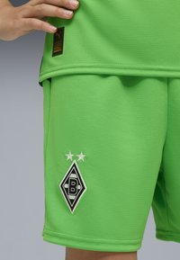 Käsi pitää kirkkaan vihreän urheilupaidin helmaa, joka on yhdistetty matching-shortsien kanssa, joissa on mustavalkoinen "B"-logo ja kaksi tähteä.