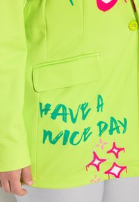 Neonvihreä blazer, jossa on tasku, ja siinä on kirkkaan sinistä ja pinkkiä tekstiä, joka sanoo "HAVE A NICE DAY" sekä tähti grafiikoita oikeassa alakulmassa.