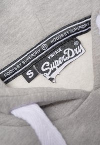 Interno della felpa grigia con una fodera morbida. Presenta un'etichetta nera con "SuperDry" in bianco, taglia "S", e dettagli del logo in bianco e nero.