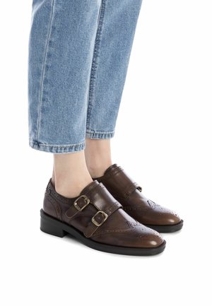 Pies que llevan zapatos de brogue de doble hebilla de cuero marrón combinados con jeans de denim azul claro cortos sobre fondo blanco.