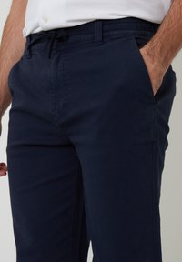 Hombre vistiendo pantalones azul marino con cintura ajustable y manos en los bolsillos, acompañado de una camisa blanca.