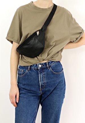 Personne portant un t-shirt marron clair et un jean bleu avec un sac bandoulière en cuir noir fermé par une fermeture éclair sur la poitrine.