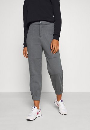 Person iført grå afkortede joggingbukser med knapdetaljer og hvide Nike-sneakers, stående foran en ensfarvet lys baggrund.