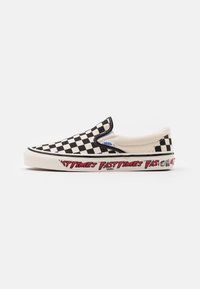 Chaussures slip-on en toile à carreaux noir et blanc, avec une semelle blanche et un texte graphique rouge sur le côté.