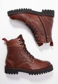 Bruna läderankelboots med texturerad yta, metallöglor, bruna snören, siddragkedja och svart gummisula med klack. Röd dragflik baktill.