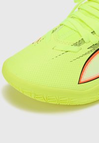 Scarpa da ginnastica di un giallo brillante con parte superiore strutturata, caratterizzata da dettagli neri e arancioni e un design a profilo basso. I lacci sono visibili.