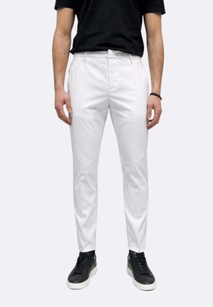 Uomo che indossa pantaloni bianchi aderenti, maglietta nera, braccialetto nero e sneakers nere con suole bianche, in piedi davanti a uno sfondo semplice.