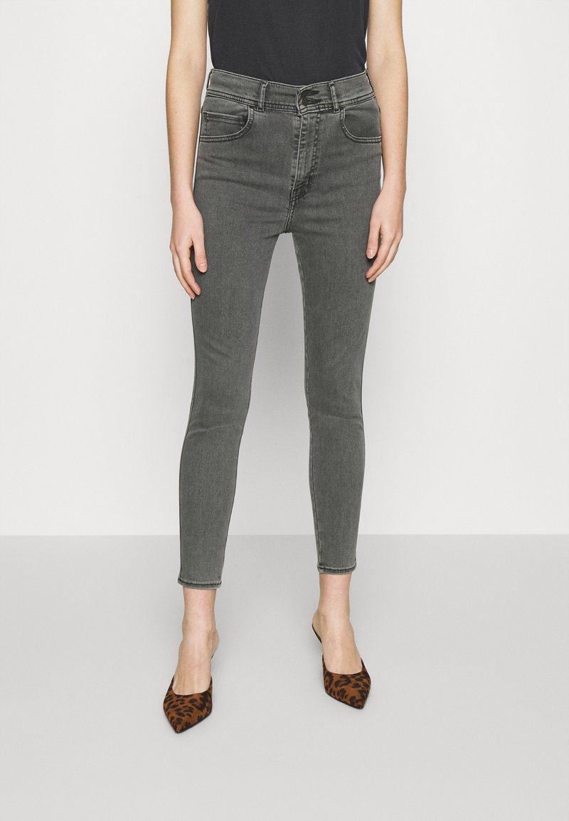 Personne portant un jean gris taille haute et slim, avec des talons pointus imprimés léopard, debout devant un fond clair uni.